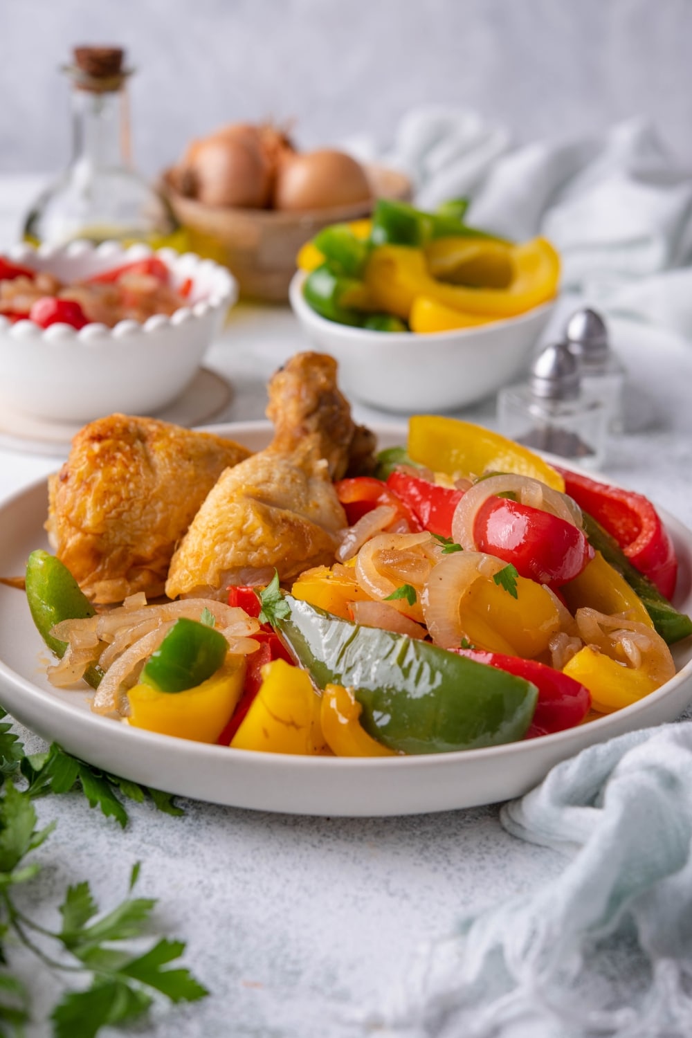 Sauteed bell peppers and onions garnished with parsley and served with fried chicken on a white plate. Behind the plate are bowls of sliced peppers and whole onions as well as a glass bottle of olive oil.