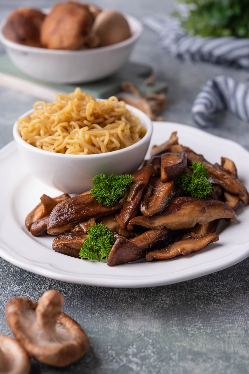 Sauteed shiitake mushrooms garnished with parsley on a white plate next to a small bowl of noodles. In the back is a bowl of raw shiitake mushrooms.