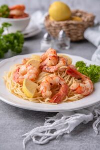 Sauteed shrimp on spaghetti noodles garnished with parsley and wedges of lemon on a white plate. In the background is a basket of lemons, salt and pepper shakers, and a bowl of more sauteed shrimp.