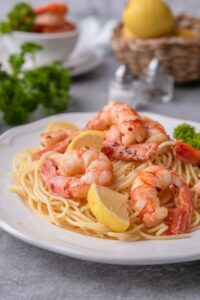 Close up of sauteed shrimp on spaghetti noodles garnished with lemon wedges and curly parsley on a white plate. In the back is a basket of lemons, salt and pepper shakers, and a bowl of more sauteed shrimp.
