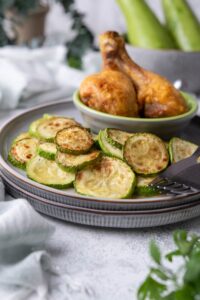 Close up of sauteed zucchini slices. They're served on a stacked pair of blue grey plates with a small bowl of two fried chicken drumsticks. Part of two forks can be seen on the plate and part of a bowl of fresh zucchinis can be seen in the back.