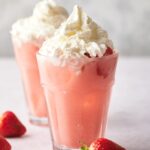 A pink drink in a glass with whipped cream on top. In front of that is a few strawberries and behind it is another drink.