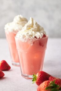 A pink drink in a glass with whipped cream on top. In front of that is a few strawberries and behind it is another drink.