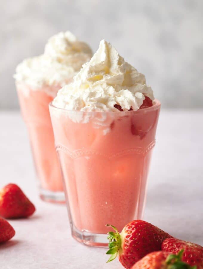 A pink drink in a glass with whipped cream on top. In front of that is a few strawberries and behind it is another drink.