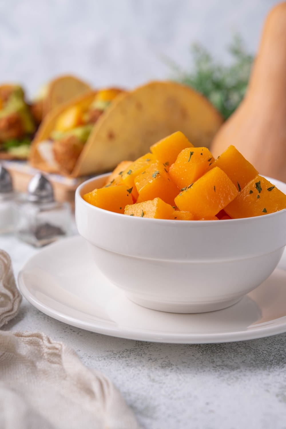 Sauteed butternut squash cubes in a white bowl set on top of a white plate. In the back is a plate of hard shell tacos with various toppings and butternut squash cubes. Next to it, part of a butternut squash can be seen.