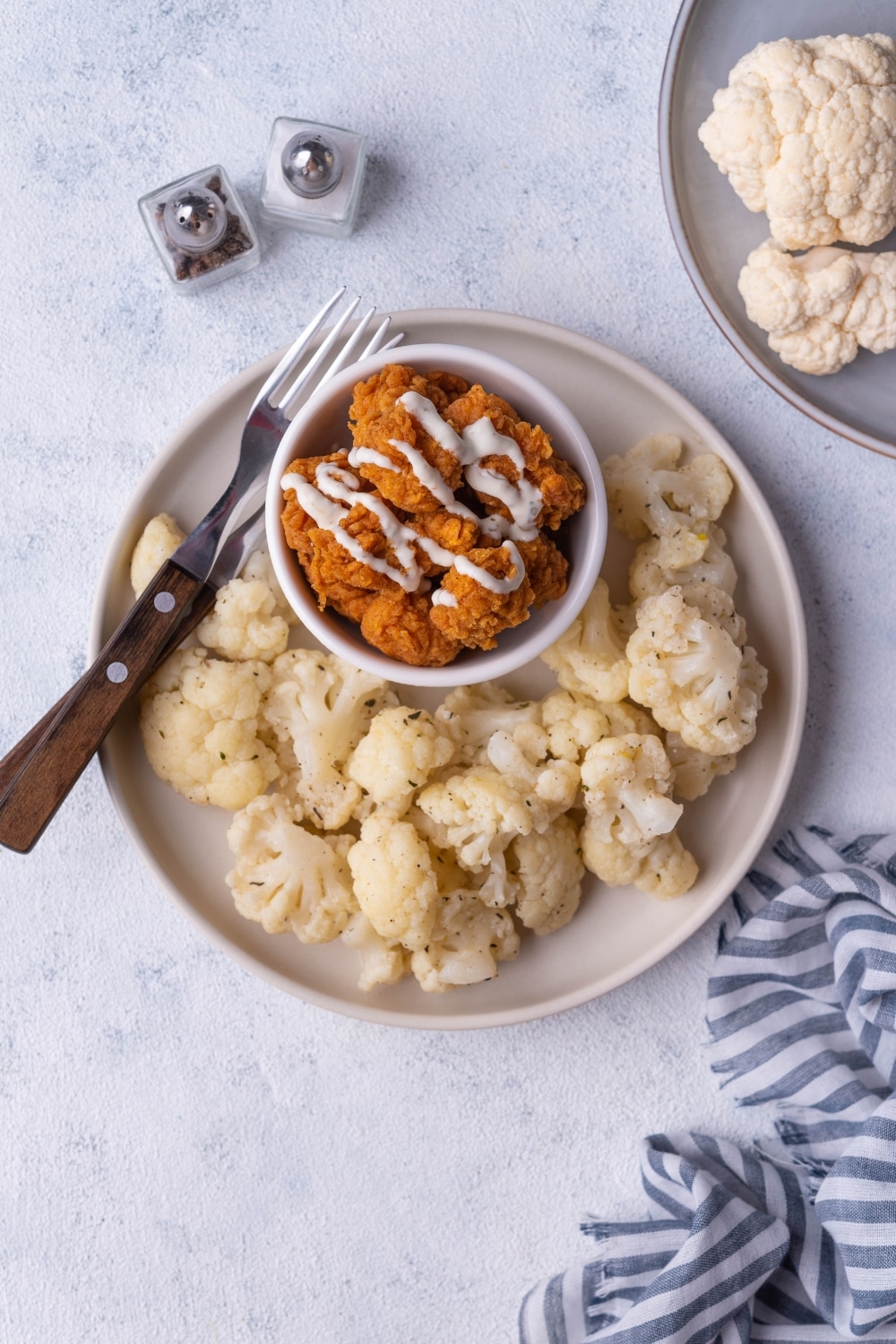 Top view of sauteed cauliflower flortes on a white plate with. On the same plate is a small bowl of fried chicken drizzled with white sauce and two forks with wooden handles. Surrounding the plate is a plate of raw cauliflower, a draped tea towel, and a pair of salt and pepper shakers.