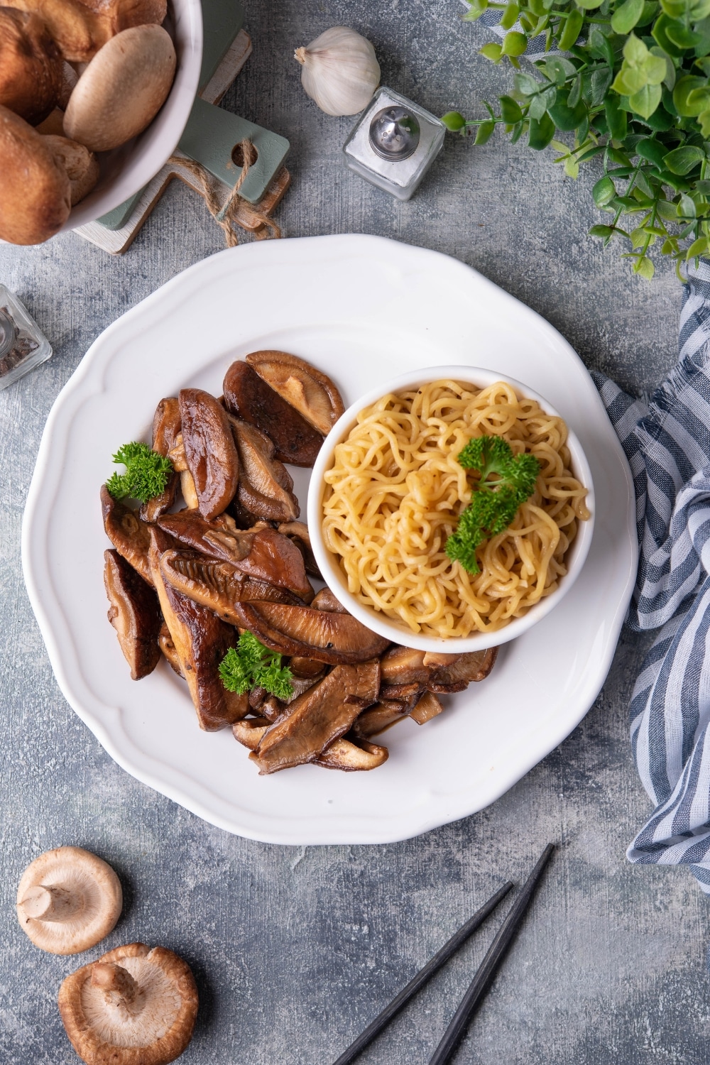 Top view of sauteed shiitake mushrooms garnished with parsley on a white plate next to a small bowl of noodles. Surrounding it is part of a bowl of raw shiitake mushrooms, fresh produce, and a pair of black chopsticks.