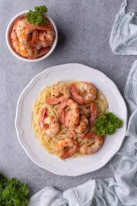 Top view of a plate of sauteed shrimp on spaghetti noodles and a bowl of sauteed shrimp. Both are garnished with a piece of curly parsley on the side.