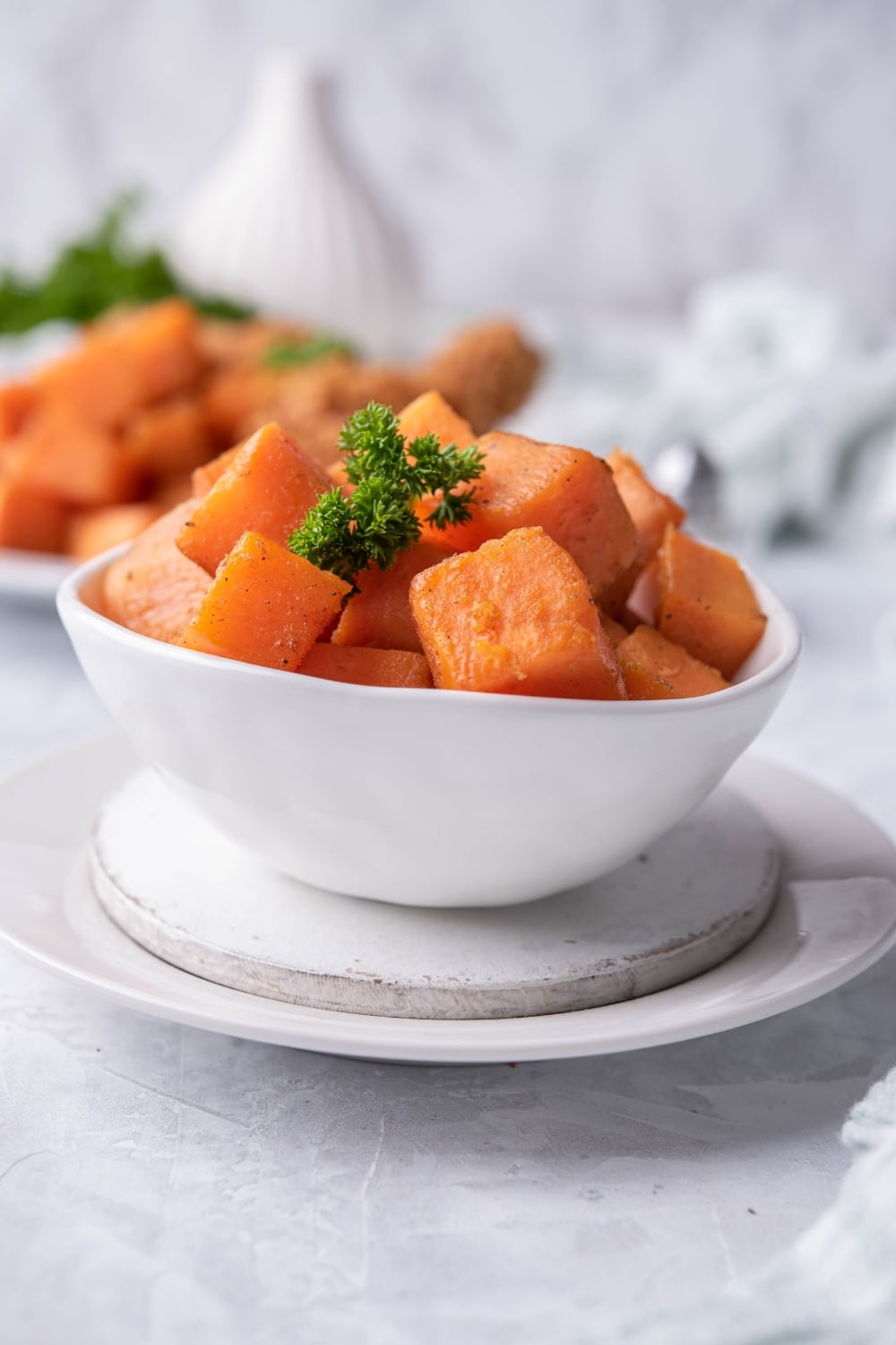 Sauteed sweet potato cubes garnished with curly parsley in a small white bowl over a white plate. Behind it is a plate of more sauteed sweet potatoes with fried chicken.