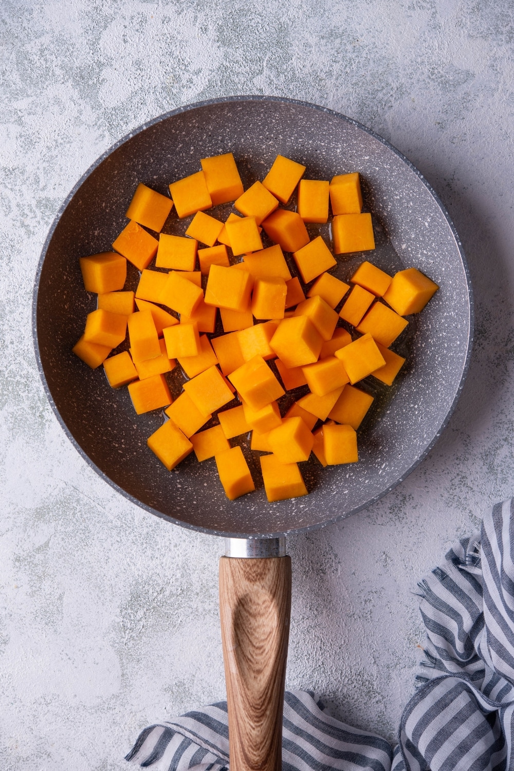 Butternut squash cubes over oilve oil in a grey speckled pan with a wooden handle. The pan is resting on a grey countertop.