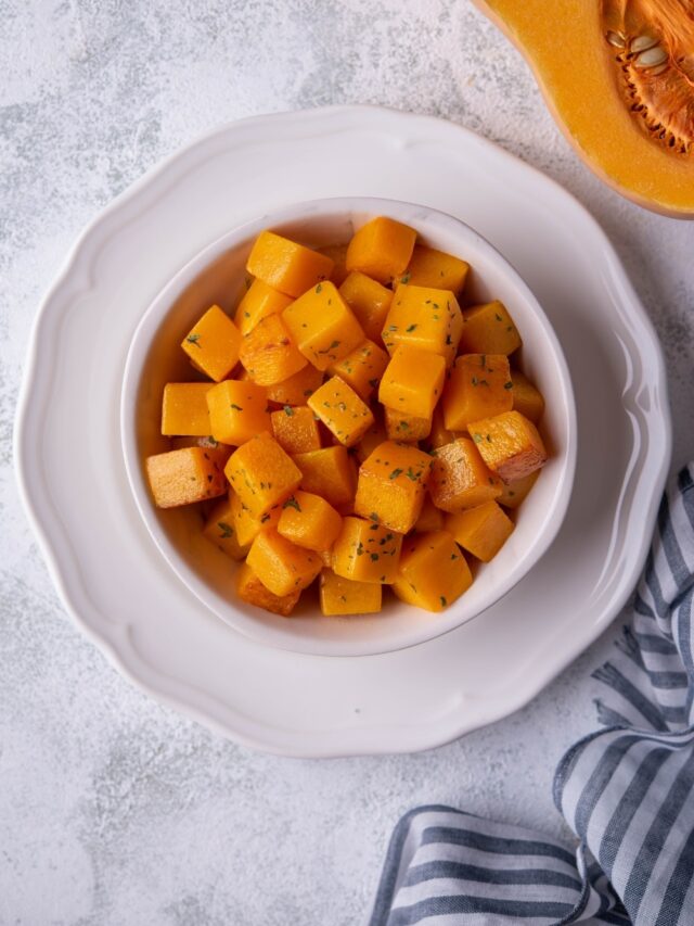 Top view of sauteed butternut squash cubes in a white bowl set on top of a white plate. In the upper corner is part of a halved butternut squash and in the lower corner is a striped tea towel.