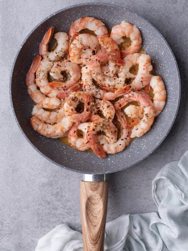 Sauteed shrimp in a grey speckled pan with a wooden handle. The shrimp are covered in chili flakes and dried herbs in a garlic butter sauce. The pan is resting on a grey countertop.
