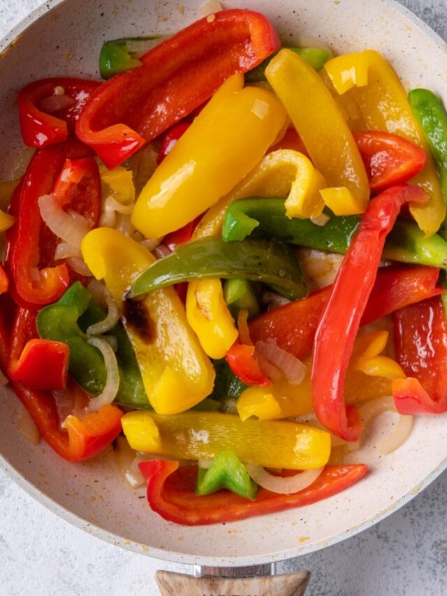 Close up of sauteed bell peppers and onions in a white speckled skillet with a wooden handle. The skillet is resting on a grey countertop.