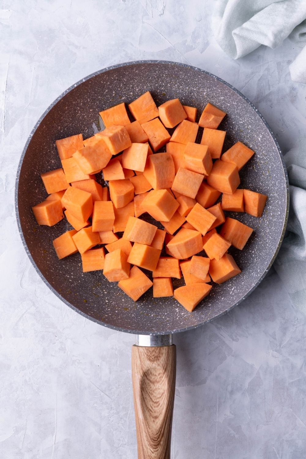 Uncooked diced sweet potatoes over oil in a grey speckled skillet with a wooden handle. The skillet is on a grey countertop next to a tea towel.