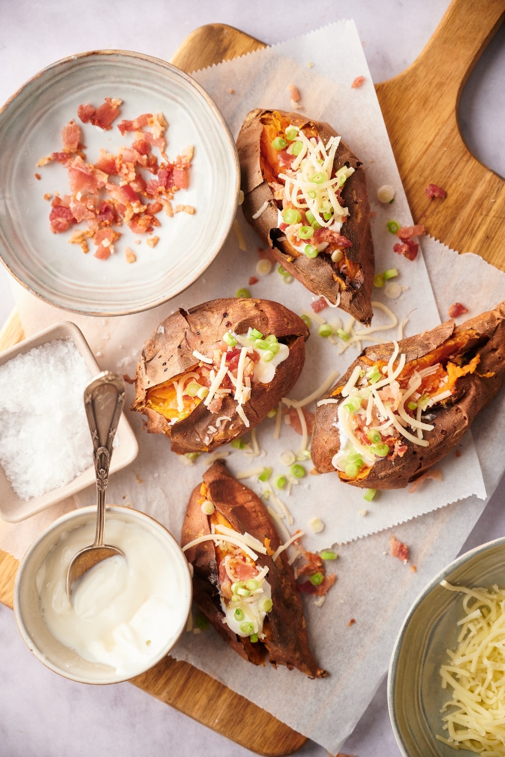 Top view of baked sweet potatoes garnished with shredded mozzarella cheese, bacon bits, sour cream, and chopped green onions on a parchmennt paper lined cutting board. Surrounding the sweet potatoes are bowls of bacon bits, sour cream, shredded mozzarella cheese, and salt.