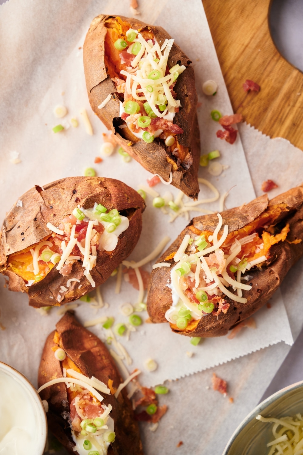 Top view of four baked sweet potatoes garnished with shredded mozzarella cheese, bacon bits, sour cream, and chopped green onions on a parchmennt paper lined wooden cutting board.