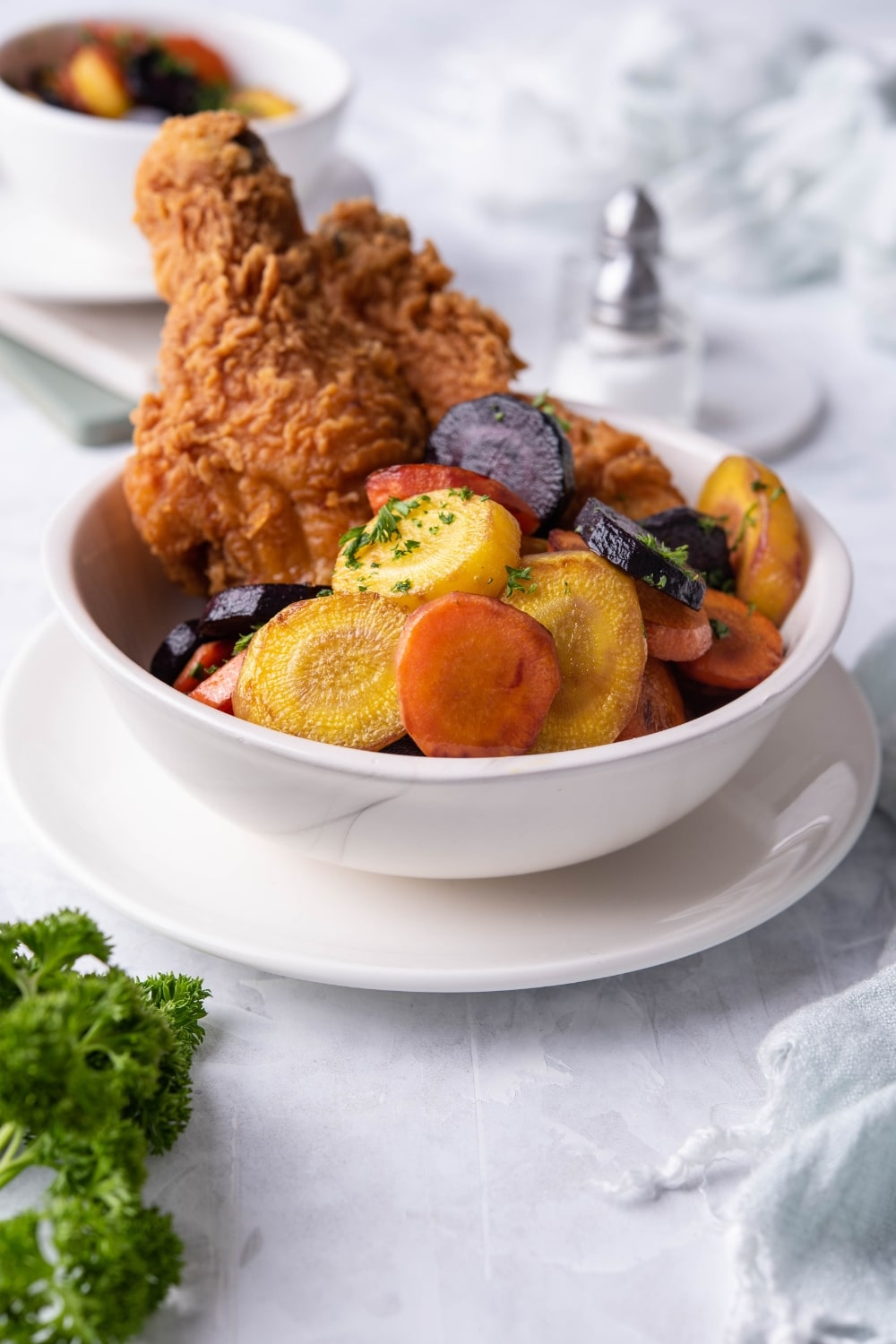 Colorful sauteed carrots garnished with chopped parsley in a white bowl with breaded fried chicken. In front of the bowl is a bunch of fresh parsley and behind it is another bowl of sauteed carrots and a pair of salt and pepper shakers.