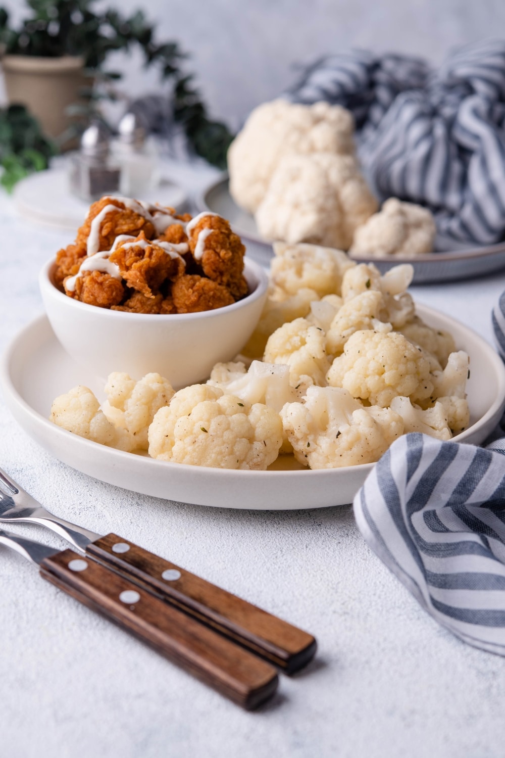 Sauteed cauliflower florets on a white plate with a small bowl of fried chicken drizzled with white sauce. Part of a pair of forks with wooden handles is in front of the plate and behind it is a plate of raw cauliflower and a pair of salt and pepper shakers.