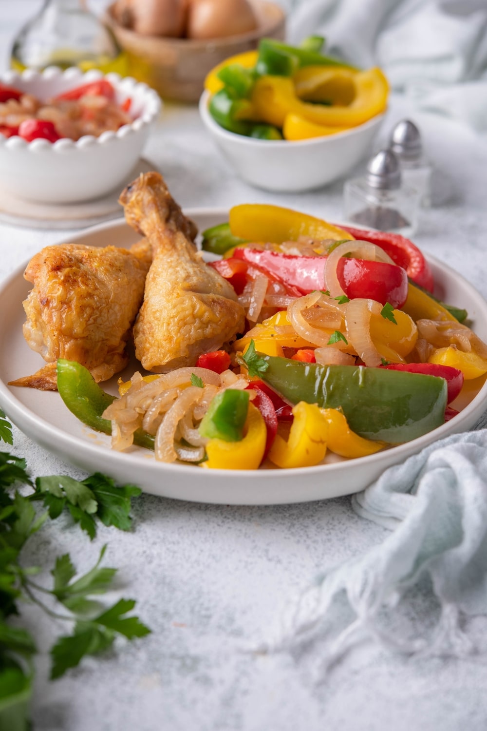 Sauteed bell peppers and onions garnish with fresh parsley and served with two pieces of fried chicken on a white plate. Behind the plate are salt and pepper shakers, bowls of whole onions, chopped peppers, and more sauteed peppers and onions, and a glass bottle of olive oil.