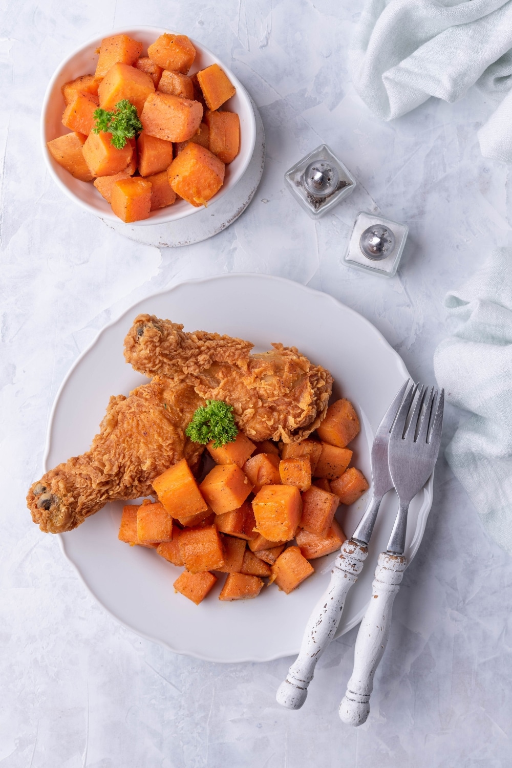 Top view of sauteed sweet potatoes on a plate with two fried chicken drumsticks and two forks with white handles. Next to the plate is a small bowl of sauteed sweet potatoes and a pair of salt and pepper shakers.