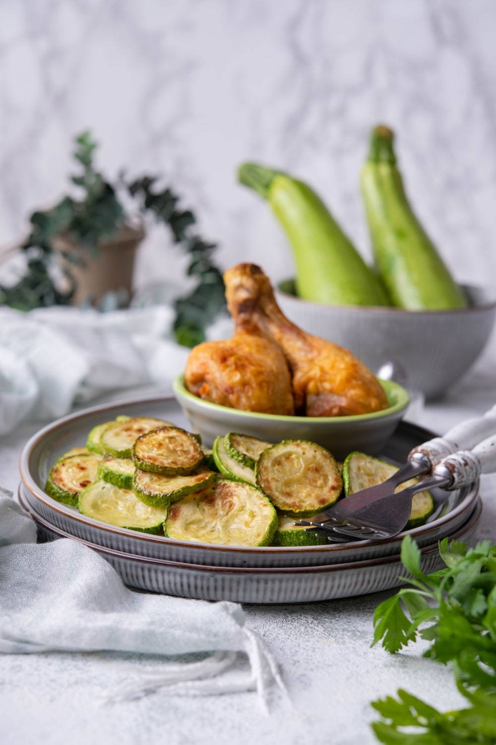 Sauteed zucchini slices on a blue grey plate with a small bowl of two fried chicken drumsticks and two forks. In the back is a bowl of fresh zucchinis.