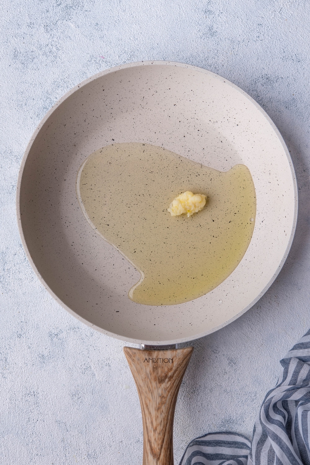 Olive oil and garlic paste in a white speckled skillet with a wooden handle. The skillet is on a grey countertop next to a striped tea towel.