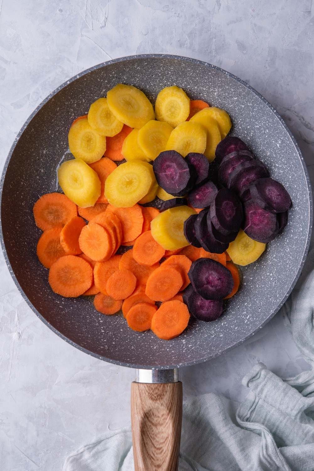 Slices of violet, yellow, and orange carrots over oil in a grey speckled skillet with a wooden handle. The skillet is on a grey countertop next to a tea towel.