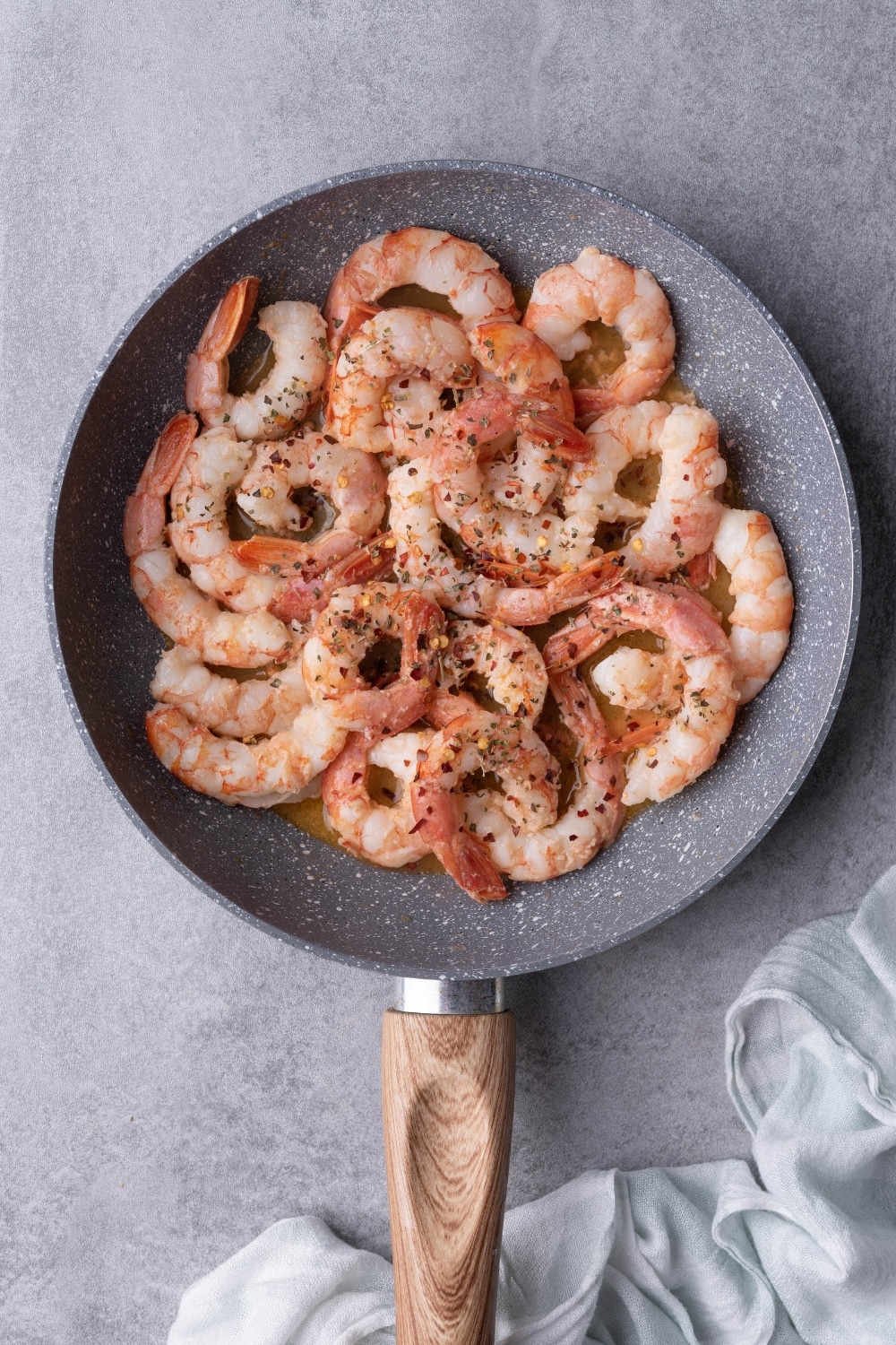 Sauteed shrimp in a grey speckled pan with a wooden handle. The shrimp are covered in chili flakes and dried herbs in a garlic butter sauce. The pan is resting on a grey countertop.