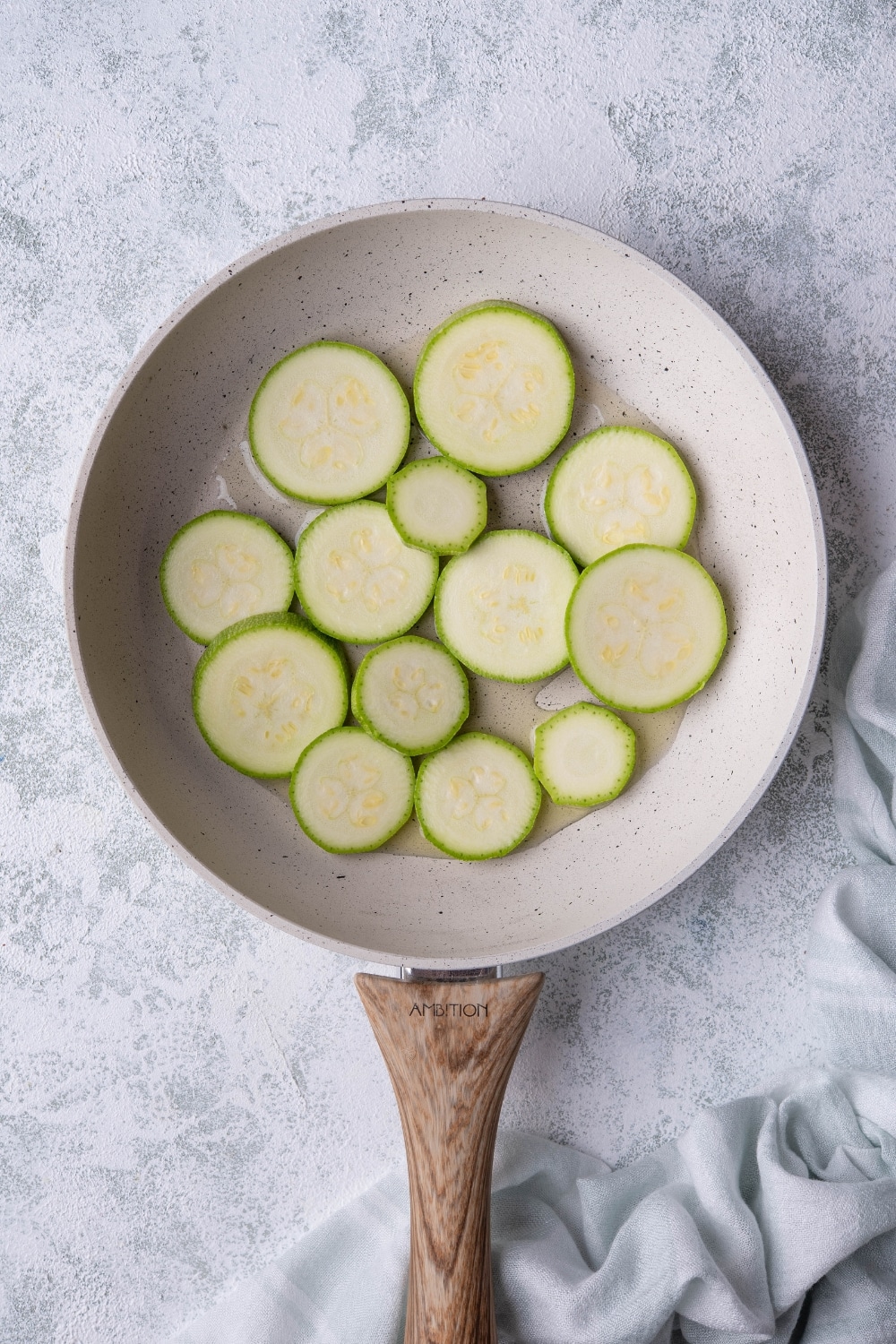 Raw zucchini slices with oil in a white speckled with a wooden handle. The pan is on a grey countertop and a tea towel is on the side.
