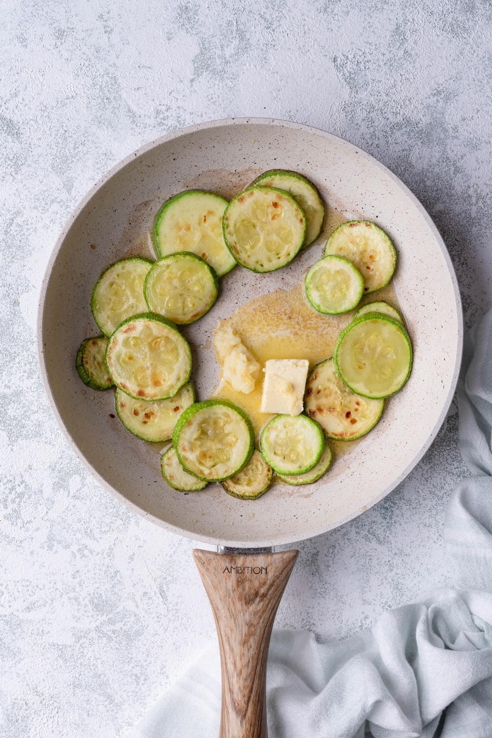 Sauteed zucchini slices arranged on the edges of a white speckled with a wooden handle. In the center of the pan is butter and garlic paste. The pan is on a grey countertop and a tea towel is on the side.