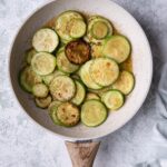 Sauteed zucchini slices with garlic butter in a white speckled with a wooden handle. The pan is on a grey countertop and a tea towel is on the side.