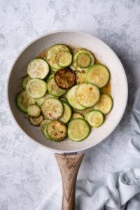 Sauteed zucchini slices with garlic butter in a white speckled with a wooden handle. The pan is on a grey countertop and a tea towel is on the side.