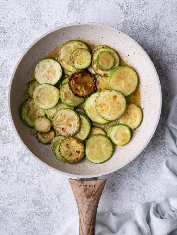 Sauteed zucchini slices with garlic butter in a white speckled with a wooden handle. The pan is on a grey countertop and a tea towel is on the side.