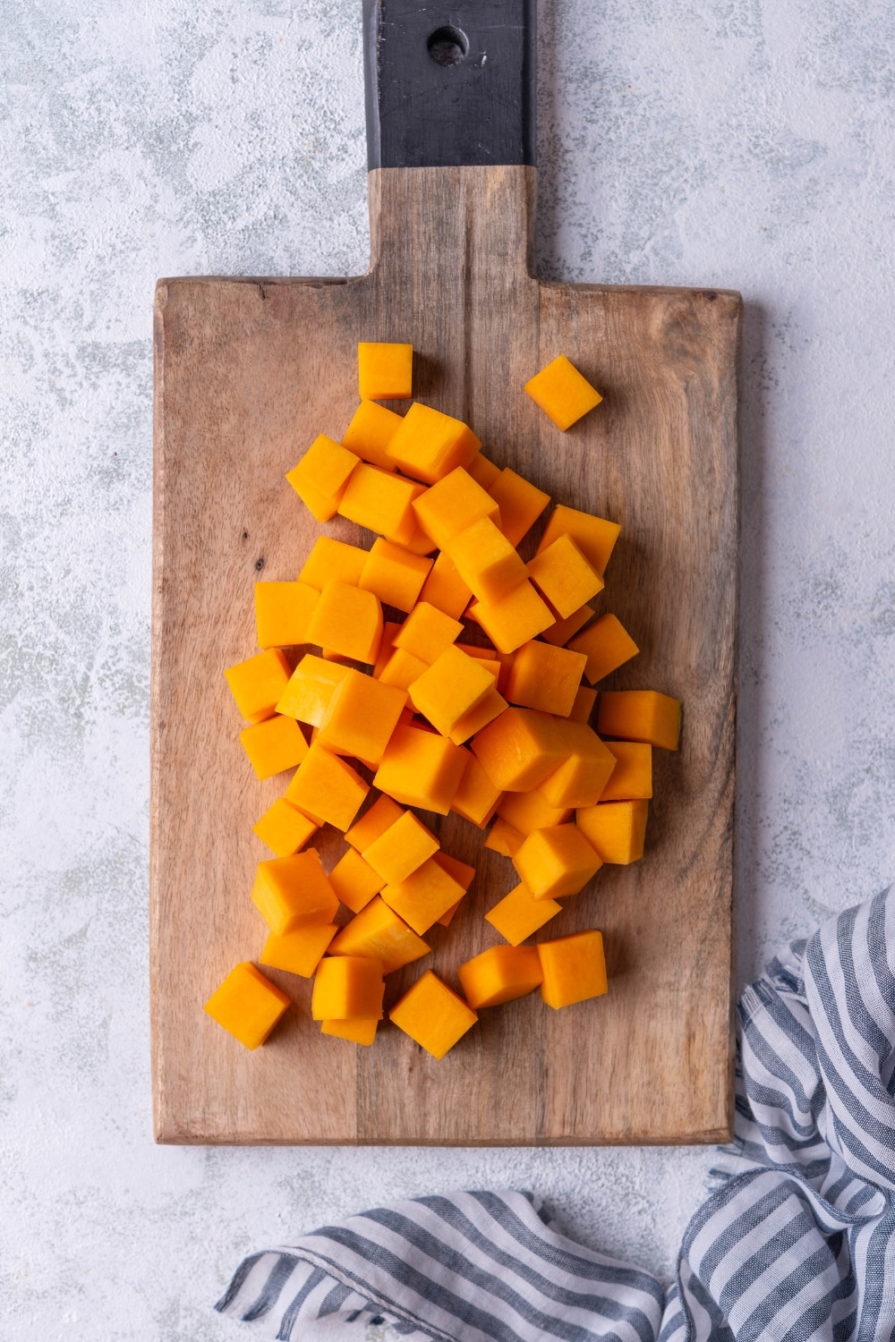 Raw butternut squash cubes on a wooden cutting board with a black handle. The cutting board is resting on a grey countertop.