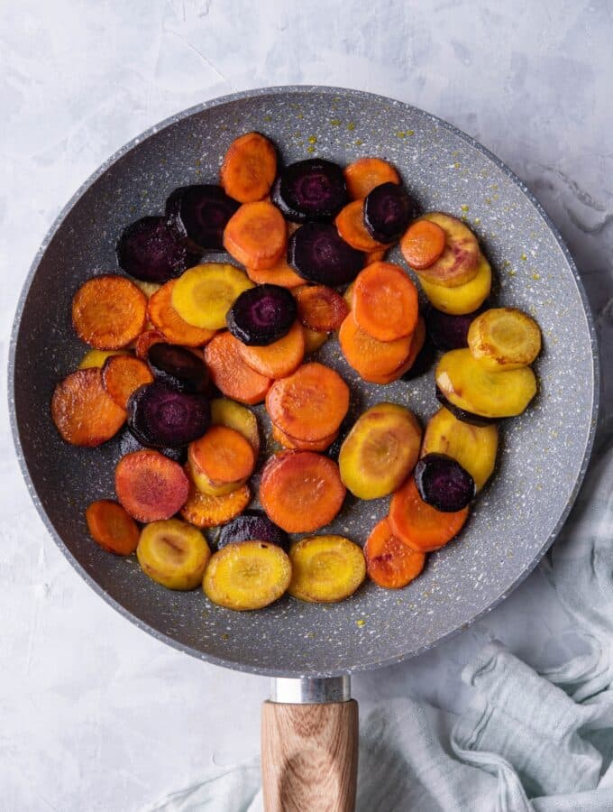 Sauteed slices of violet, yellow, and orange carrots in a grey speckled skillet with a wooden handle. The skillet is on a grey countertop next to a tea towel.