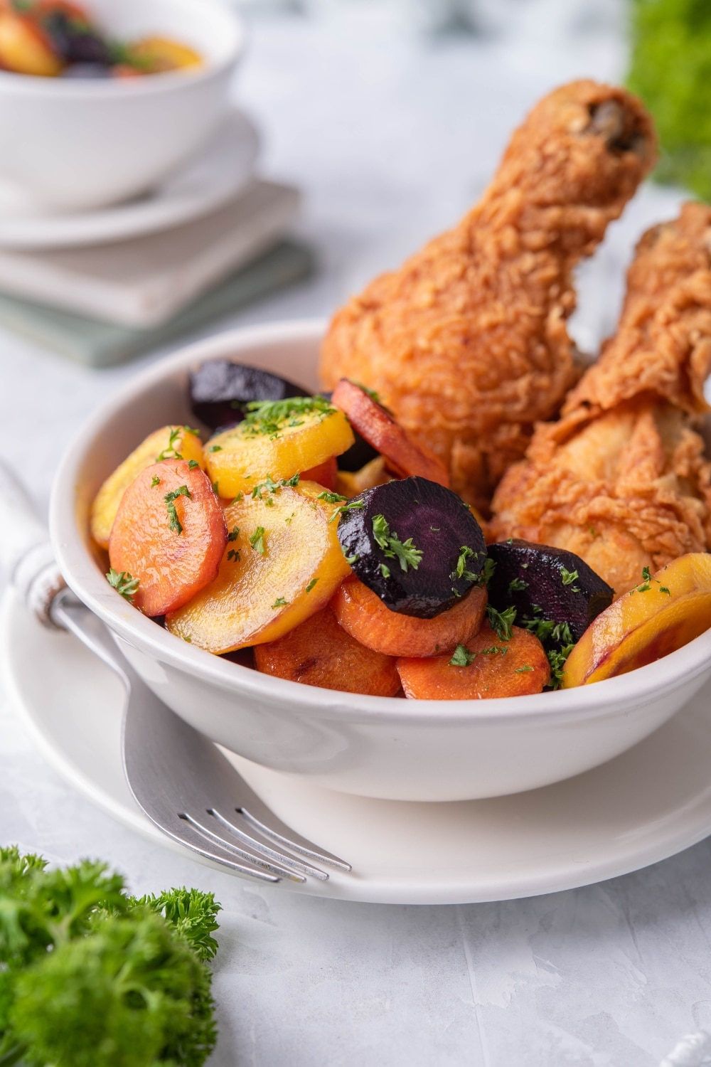 Close up of sauteed carrots garnished with chopped parsley. The carrots are in a white bowl next to a pair of fried chicken drumsticks. Part of another bowl of sauteed carrots can be seen in the back.