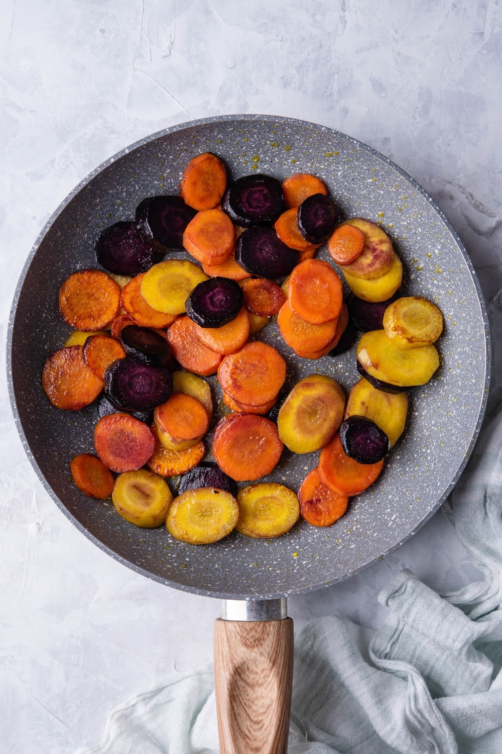 Sauteed slices of violet, yellow, and orange carrots in a grey speckled skillet with a wooden handle. The skillet is on a grey countertop next to a tea towel.