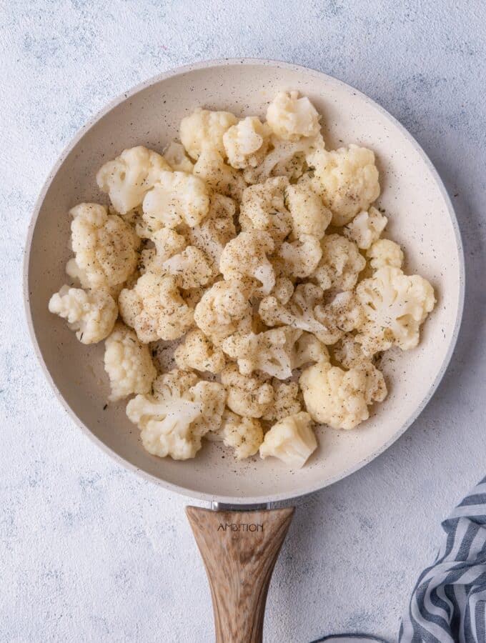 Seasoned cauliflower florets in a white speckled skillet with a wooden handle. The skillet is on a grey countertop next to a striped tea towel.