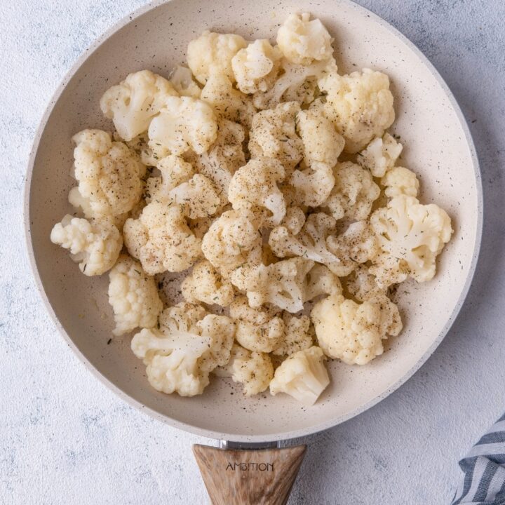 Seasoned cauliflower florets in a white speckled skillet with a wooden handle. The skillet is on a grey countertop next to a striped tea towel.