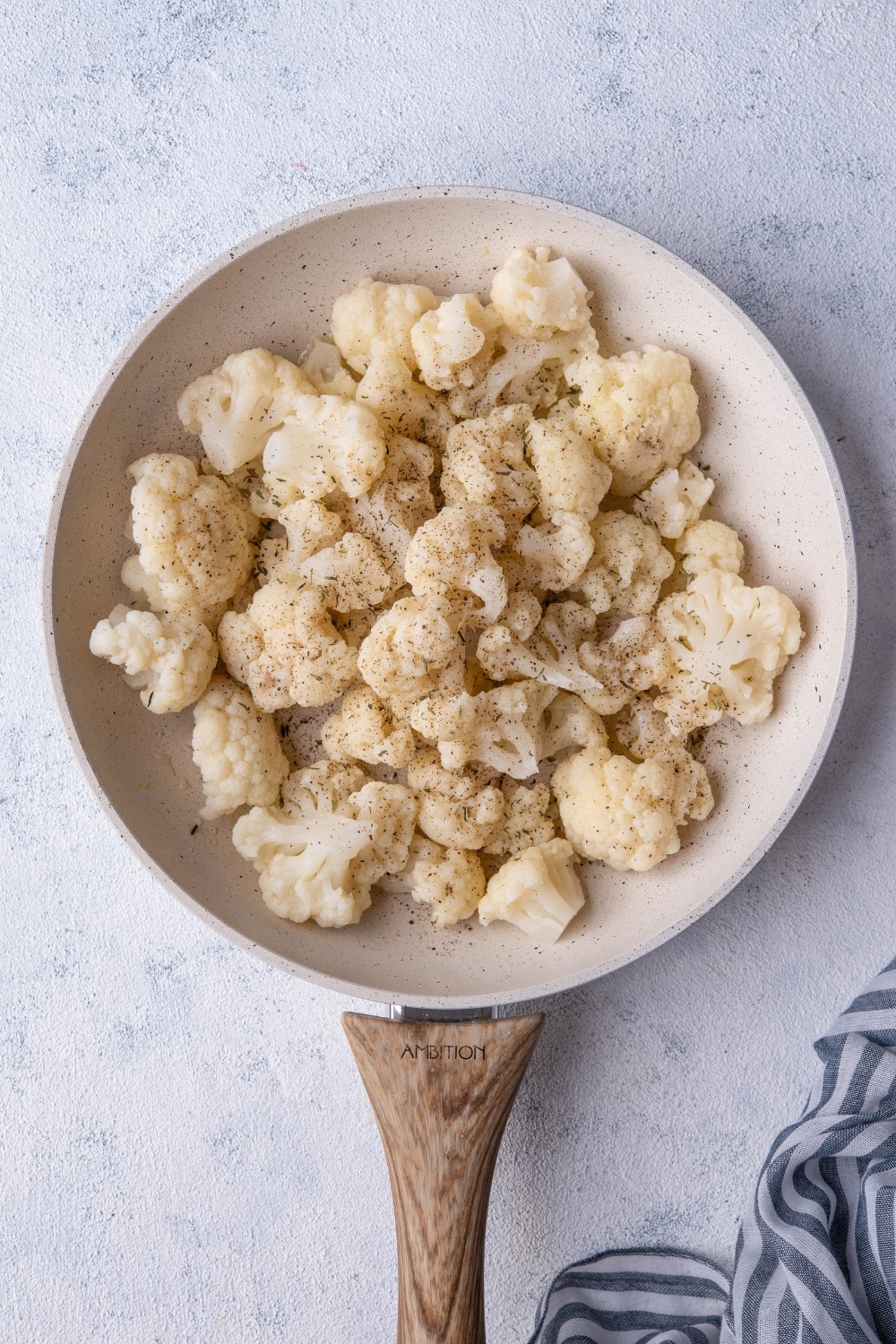 Seasoned cauliflower florets in a white speckled skillet with a wooden handle. The skillet is on a grey countertop next to a striped tea towel.