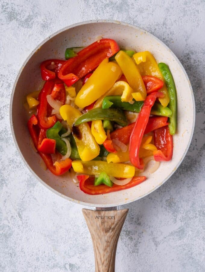 Sauteed bell peppers and onions in a white speckled skillet with a wooden handle. The skillet is on a grey countertop.