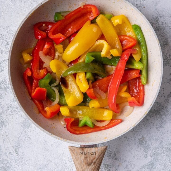 Sauteed bell peppers and onions in a white speckled skillet with a wooden handle. The skillet is on a grey countertop.