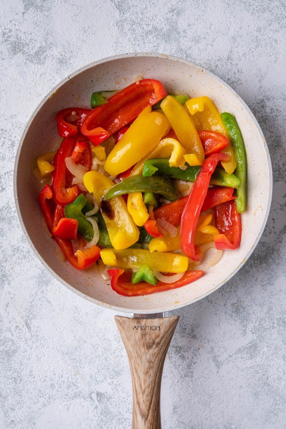 Sauteed bell peppers and onions in a white speckled skillet with a wooden handle. The skillet is on a grey countertop.