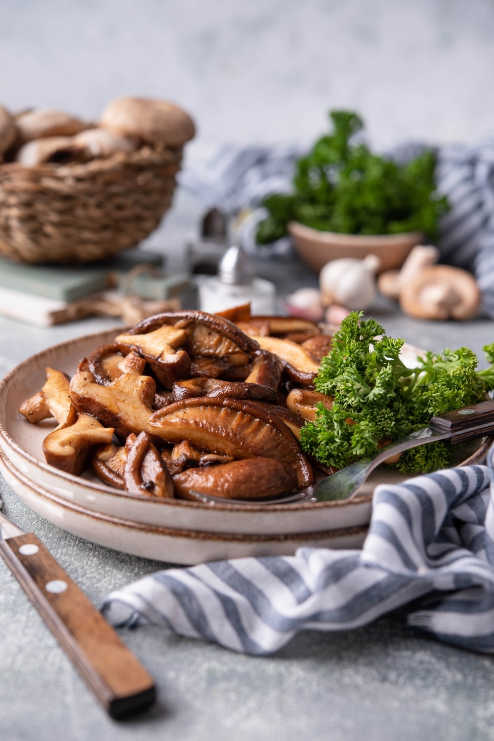 Sauteed shiitake mushrooms with curly parsley on stacked beige plates with a fork and knife. In the back is a basket of fresh mushrooms and a small bowl of parsley.