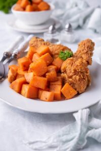 Sauteed sweet potatoes on a plate with two breaded chicken drumsticks. Two forks with white handles are on the plate and behind is a pair of salt and pepper shakers and a small bowl of sauteed sweet potatoes.