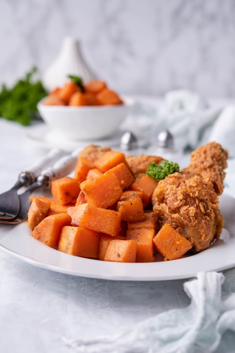 Sauteed sweet potatoes on a plate served with fried chicken drumsticks. Two forks with white handles are on the plate and behind is a small bowl with more sauteed sweet potato cubes and a pair of salt and pepper shakers.