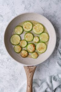 Sauteed zucchini slices with oil in a white speckled with a wooden handle. The pan is on a grey countertop and a tea towel is on the side.