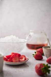 A plate of sliced strawberries, a bowl of ice, and a pitcher of tea on a counter.