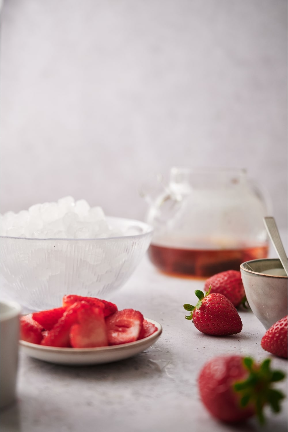 A plate of sliced strawberries, a bowl of ice, and a pitcher of tea on a counter.