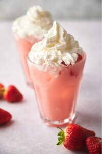 Whipped cream on top of a creamy pink drink in a glass. Behind it is another drink on the same grey counter.